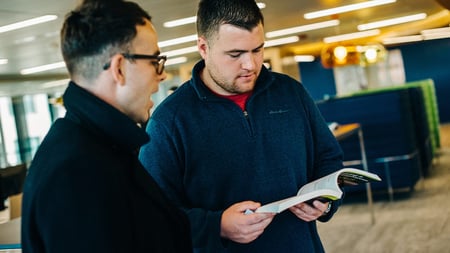 two-male-employees-talking-reading-book